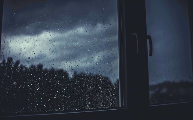 A close up scene of a window with raindrops on the window pane overlooking a dark and gloomy sky with dense clouds.