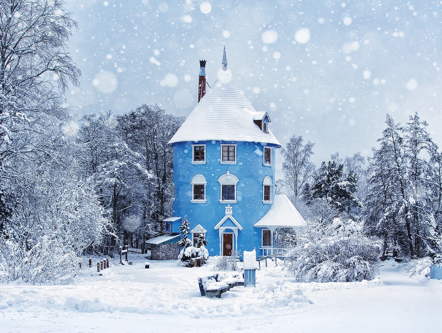 In the midst of a snowstorm stands a long and blue cylinder shaped house with three floors surrounded by inches of snow and forest trees.